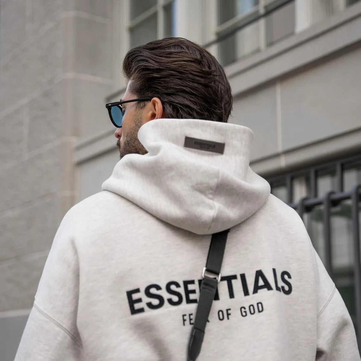 Person wearing a white hoodie with 'ESSENTIALS FEAT OF GOD' text, standing in front of a building.